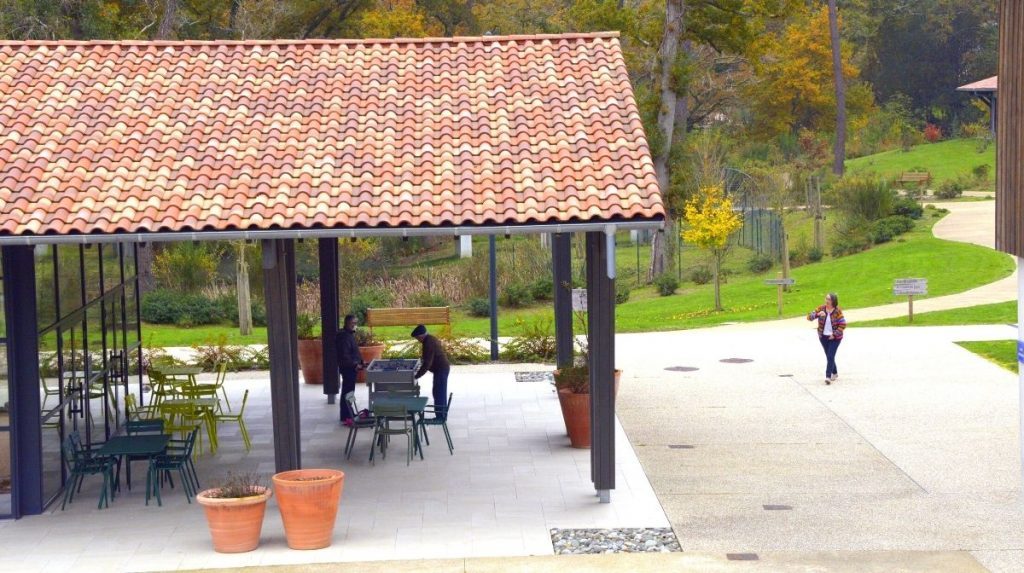 Photographie de la place du village landais, avec un préau équipé de tables et de chaises, dans un environnement verdoyant.