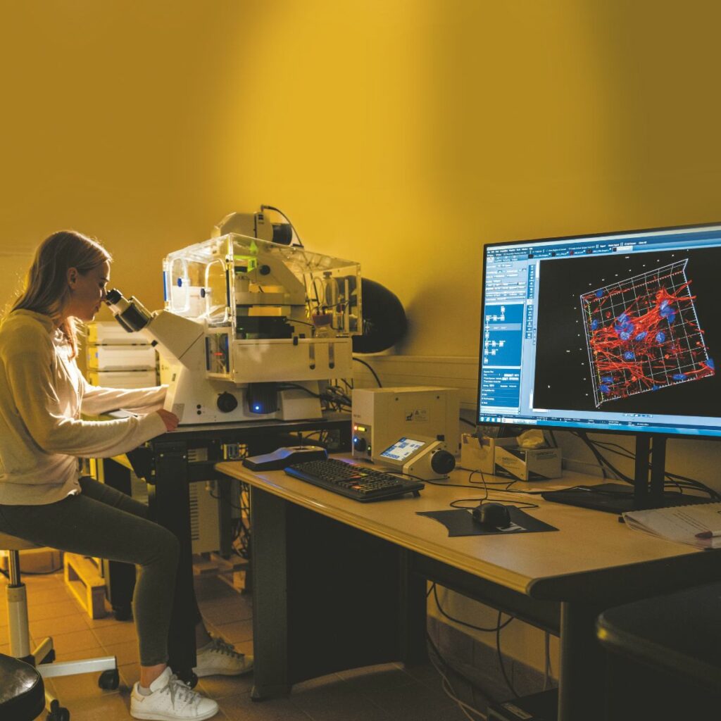 Dans un laboratoire, une jeune femme réalise une observation à l’aide d’un microscope imposant. Sur une table à côté d’elle, un écran d’ordinateur présente une image qui correspond à la reconstitution en trois dimensions d’un tissu biologique.