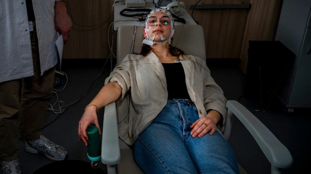 Une jeune femme est installée dans un fauteuil confortable adapté à la sieste, dans un laboratoire. Elle porte un bonnet avec des électrodes. La pièce est plongée dans la pénombre.