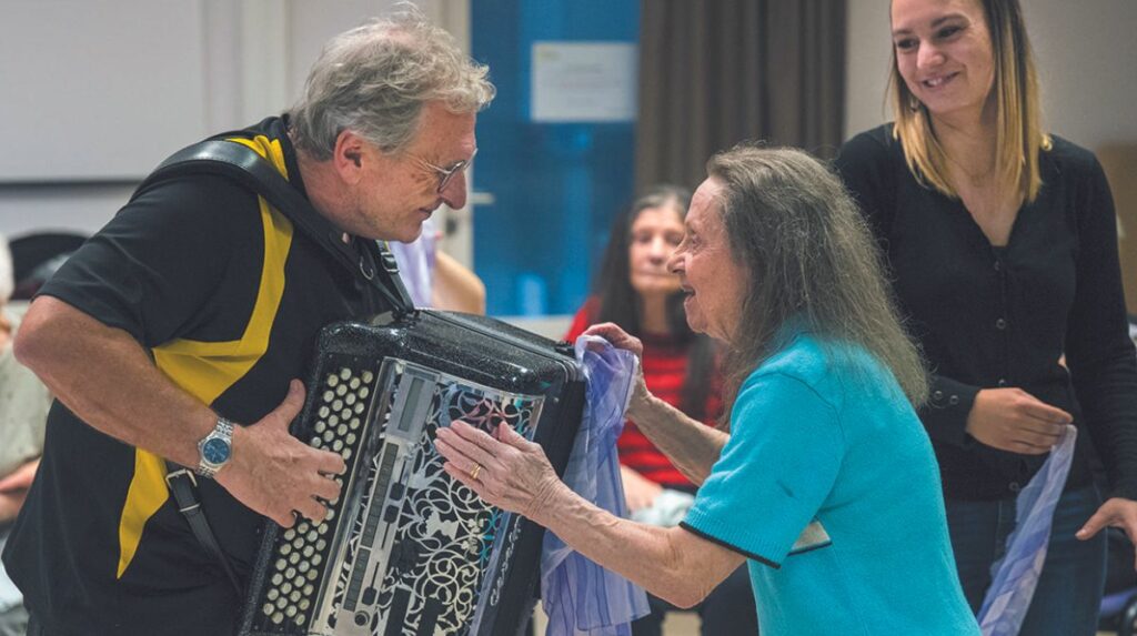 Un homme joue de l’accordéon et une femme âgée lui fait face l’air enjoué. Une jeune femme les regarde en souriant. 