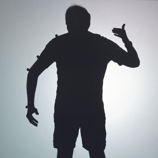 La silhouette du même homme que sur la photo précédente, en contre-jour, se détache en noir sur un fond blanc. On voit des objets sphériques de la taille de noix, fixés sur son poignet, son bras et son épaule droite. 
